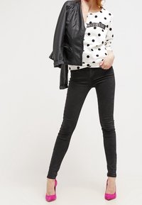 Kobieta ubrana w biały sweter w białe kropki z tekstem "NEED IS LOVE", czarne dopasowane jeansy, jaskrawo różowe szpilki oraz czarną skórzaną kurtkę zarzuconą na jedno ramię.