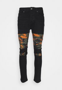 Jeans de mezclilla negra con paneles de camuflaje en naranja y verde, con detalles rasgados y un diseño de corte ajustado.