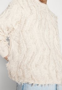 Kermaavärinen neulepaita, jossa on teksturoitu, kulunut muotoilu, pyöreä kaula-aukko ja pitkät hihat, löysällä mitoituksella.