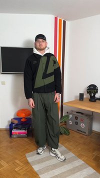 Črno in olivno zelena jakna z geometričnim vzorcem, kombinirana z ohlapnimi olivno zelenimi hlačami in sivimi supergami. Notranji ambient z dekoracijo.