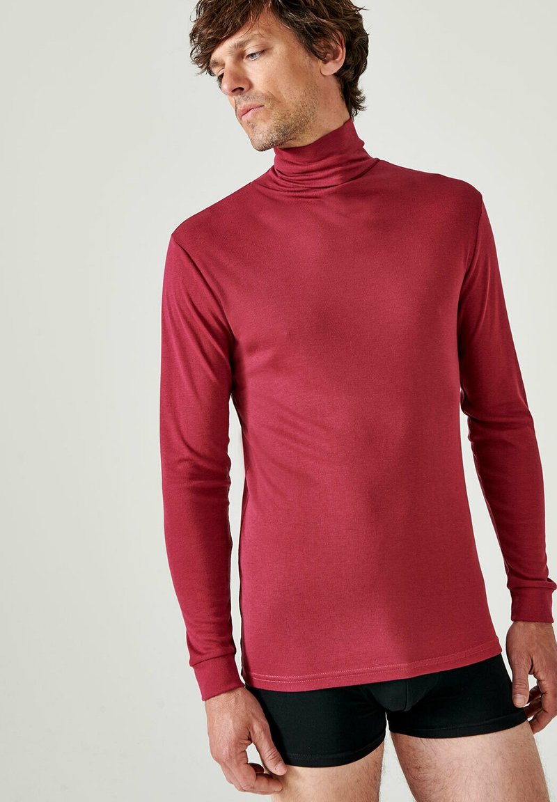 Maglietta rossa a maniche lunghe con colletto a imbuto realizzata in un tessuto liscio e elastico, con un design aderente e polsini a coste. Pantaloni corti neri sotto.