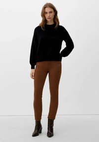 Frau steht vor weißem Hintergrund und trägt einen schwarzen Langarmpullover, braune schmal geschnittene Hose und dunkelbraune Stiefeletten.