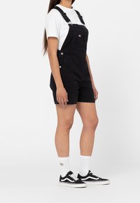 Černé denimsové kombinézy s nastavitelnými ramínky, s přední kapsou a kovovými doplňky, zkombinované s bílým tričkem a černými teniskami.