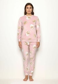 Mujer vistiendo pijamas de felpa rosa con caras de oso de peluche, de pie descalza contra un fondo liso de color claro.