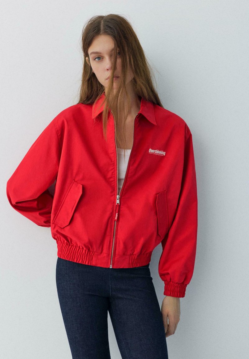 Mlajša ženska z dolgimi lasmi nosi rdečo jakno z zadrgo, na kateri piše "Preveč razmišljanja", in temno modre kavbojke, stoji ob preprosti svetli steni.