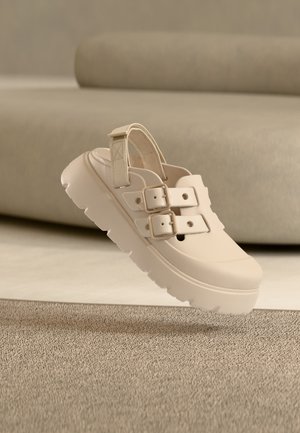 Beige chunky sole sandal with two buckle straps and a back strap, displayed on a neutral carpeted floor in front of a beige sofa.