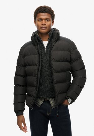 Chaqueta acolchada negra con cuello alto, diseño acolchado y puños de canalé. Llevada sobre un suéter oscuro y una camisa de cuadros, combinada con jeans oscuros.
