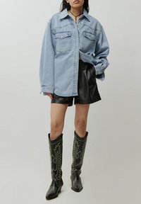 Lichtblauwe denim oversized shirt met twee opgestikte zakken aan de voorkant, gecombineerd met zwarte leren shorts en zwarte gepatternde cowboylaarzen.