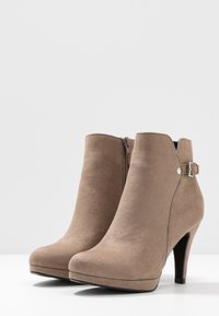 Botines de ante marrón con tacón alto, cremallera lateral y una correa de acento con una hebilla de metal. Textura suave y diseño de punta redondeada.
