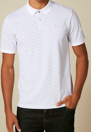 Homme portant un polo blanc à manches courtes avec de fines rayures horizontales noires et un jean foncé, la main dans la poche.