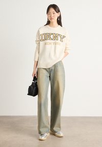 Kremfarget strikkegenser med "DKNY NEW YORK" logo, kombinert med lyseblå vide jeans. Tilbehør inkluderer en liten svart veske og sølvfargede sko.