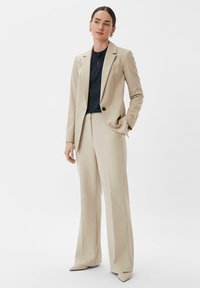 Beige maatpak met een aansluitende blazer en wijde broekspijpen. De blazer heeft een enkele zwarte knoop, gecombineerd met een donkerblauw topje eronder.