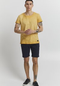 Gelbes strukturiertes T-Shirt mit umgeschlagenen Ärmeln, kombiniert mit dunkelblauen Shorts und schwarzen Sneakers. Sichtbare Tattoos an den Armen.