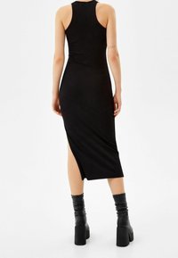 Robe midi noire côtelée avec un dos décolleté, présentant une fente latérale. Associée à des bottes noires à talons montant jusqu'aux genoux. Texture lisse et coupe ajustée.
