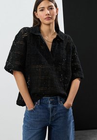 Schwarze Spitzenbluse mit Kragen und kurzen Ärmeln, die ein Karomuster aufweist. Kombiniert mit blauen Jeans.