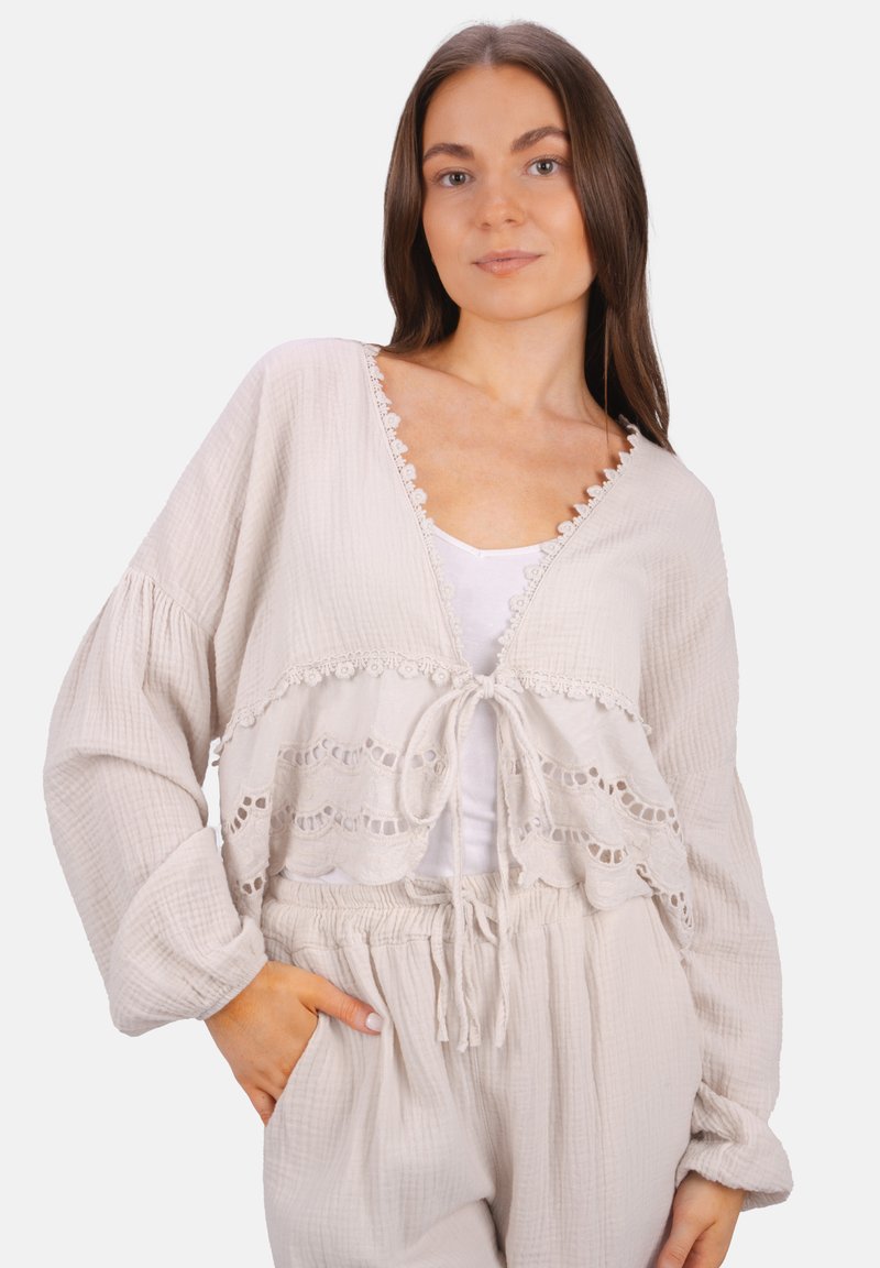 Helle beige, kurz geschnittene Strickjacke mit Spitzenbesatz, Bindeschluss und langen Ärmeln. Strukturierter Stoff und geschichtetes Design, kombiniert mit einem weißen Oberteil.