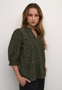 Mørkegrøn bluse i blondestof med V-udskæring, blomstermønster og pufærmer. Tekstureret stof med bølgede kanter ved ærmerne.