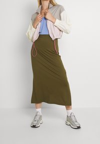 Donna che indossa una giacca cropped pastello, un top blu, una gonna midi verde oliva, calze bianche e sneakers argentate, in piedi davanti a uno sfondo semplice.