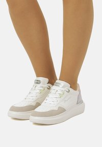 Baskets blanches avec une tige texturée, des accents en daim gris clair et des détails en pastel violet et rose. Équipées de lacets blancs et d'un logo.