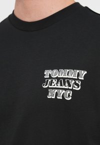 Czarny bawełniany t-shirt z okrągłym dekoltem. Na przodzie znajduje się białe logo tekstowe "TOMMY JEANS NYC" w różnych rozmiarach i stylach czcionki.