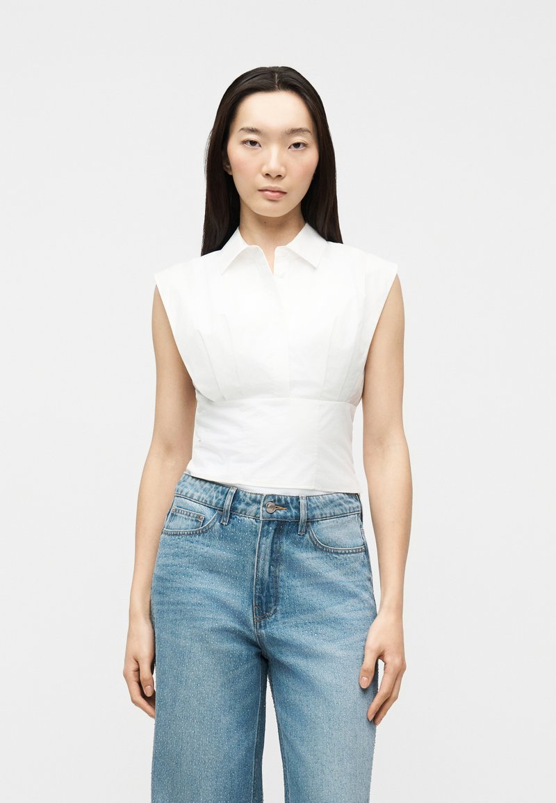 Νεαρή γυναίκα με μακριά σκούρα μαλλιά, που φοράει λευκό αμάνικο τοπ με γιακά και ψηλόμεσα ανοιχτό μπλε τζιν, σε απλό φόντο.