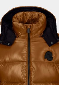 Giacca a vento marrone con colletto alto, texture imbottita, dettagli neri, zip frontale e patch rotondo del logo sul lato sinistro.