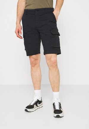 Man draagt zwarte cargoshorts, witte sokken en zwart-witte Nike sneakers, staande met één hand in de zak.