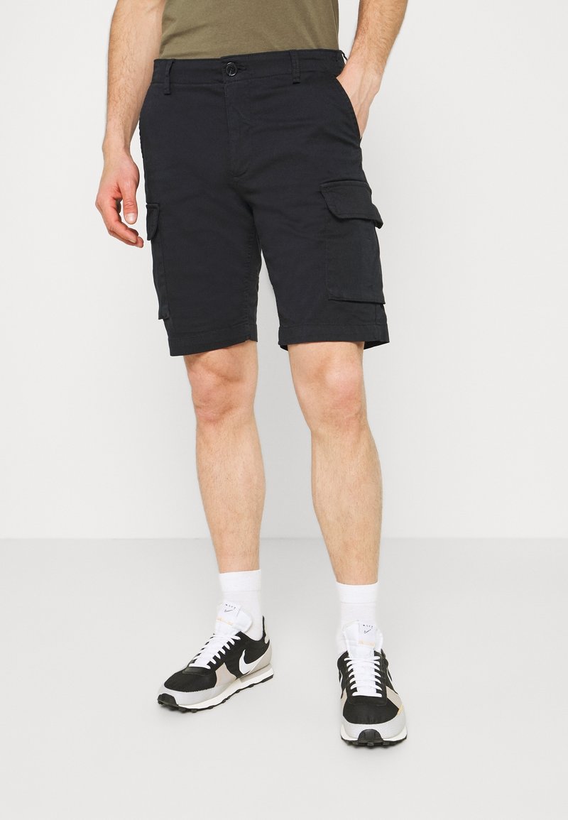 Homme portant un short cargo noir, des chaussettes blanches et des baskets Nike noires et blanches, debout avec une main dans la poche.