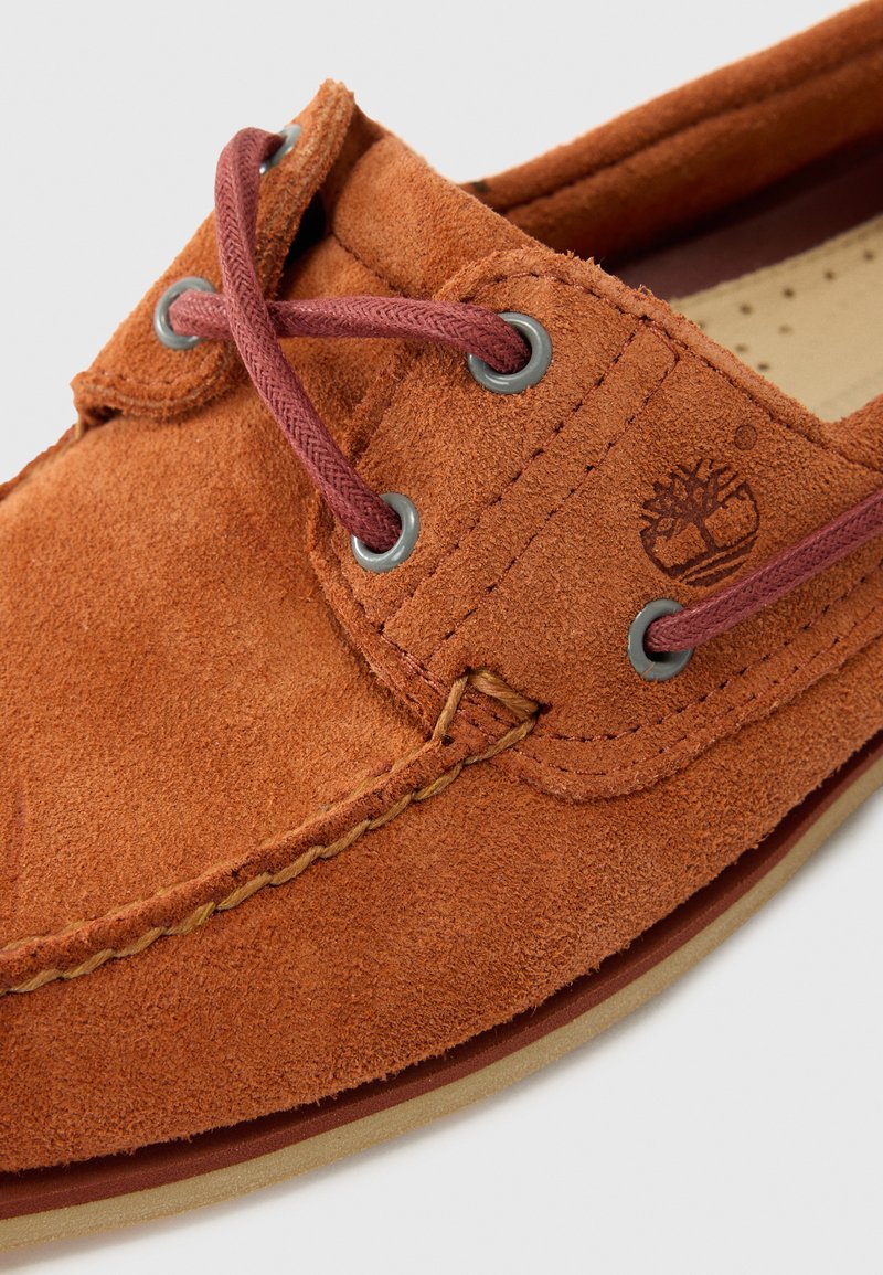 Zapato de ante marrón con un diseño cosido, que presenta cordones burdeos y ojales grises. Un logo está grabado en el lado. Suela lisa visible.