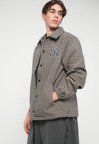 Jachetă cu carouri maronii și bej, cu guler, având un emblema de marcă pe piept, două buzunare laterale și închidere cu nasturi.