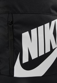 Sac à dos en tissu noir avec un grand logo Nike blanc, texture lisse et une poche zippée pour le rangement.