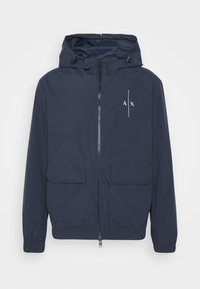 Námořnická modrá lehká bunda s kapucí, má přední zip, dvě velké kapsy a bílé logo "AX" na levém prsu.
