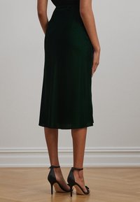 Green satin midi skirt with a smooth texture, fitted waist, and flared hem. Paired with black high-heeled sandals featuring ankle straps.