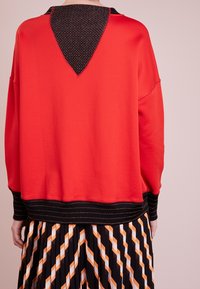 Sudadera roja con un corte en V negro texturizado en la parte posterior, con puños y dobladillo a rayas negras. Superpuesta sobre una falda plisada con patrones geométricos.