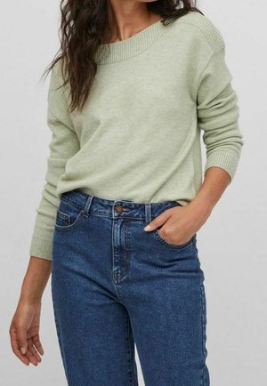 Femme portant un pull vert clair à manches longues et un jean bleu taille haute, une main dans la poche, sur un fond uni.