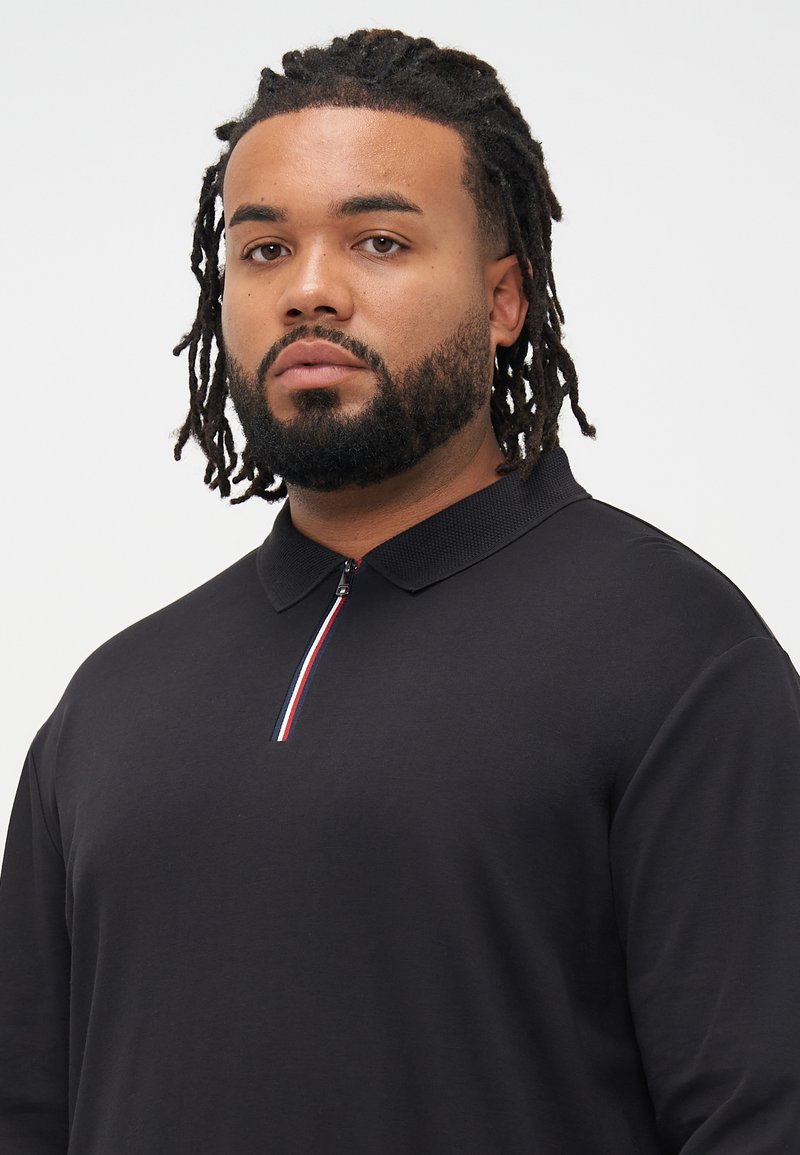 Schwarzes Poloshirt mit kurzem Reißverschlusskragen, ausgestattet mit einem Akzentstreifen in Rot, Weiß und Blau. Weicher, glatter Stoff mit normaler Passform.