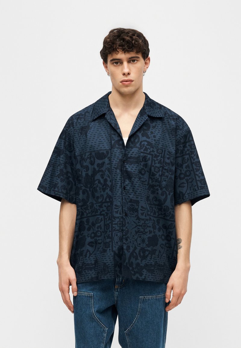 Giovane uomo con capelli ricci indossa una camicia a maniche corte con bottoni color blu scuro a motivo e jeans blu, in piedi contro uno sfondo semplice.