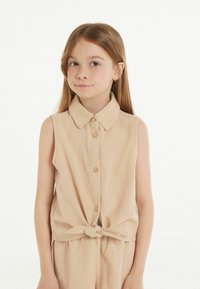 Mouwloos lichtbeige top met een kraag, knoopsluiting aan de voorkant en een strikdetail in de taille. Glad materiaal en een casual ontwerp.