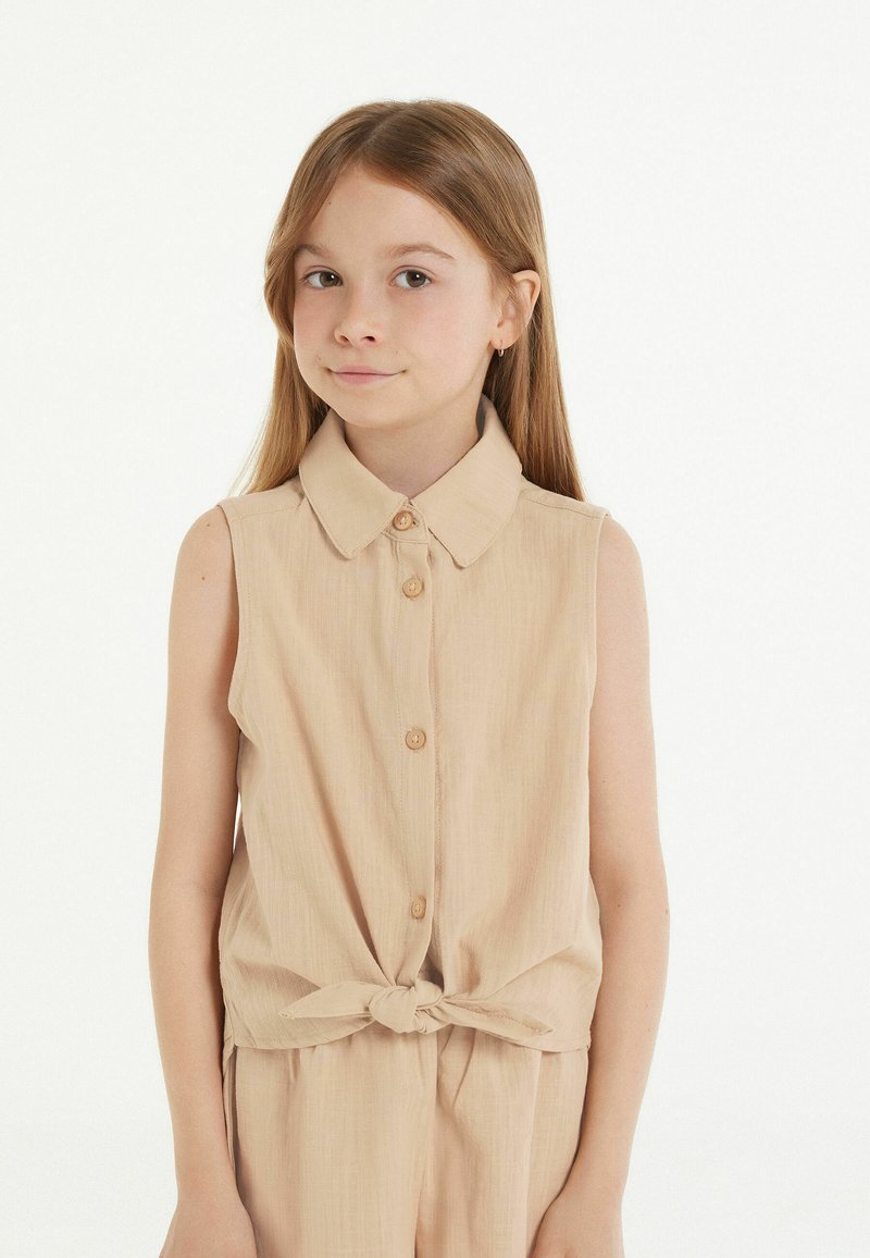 Mouwloos lichtbeige top met een kraag, knoopsluiting aan de voorkant en een strikdetail in de taille. Glad materiaal en een casual ontwerp.