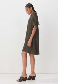 Dunkelolivgrünes, locker sitzendes Kleid mit kurzen Ärmeln, leichtem Stoff und gerader Silhouette. Tragen mit schwarzen, durchsichtigen Absatzmules.