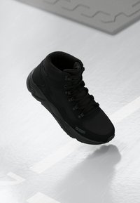 Fekete magasszárú sneaker, matt szintetikus anyagból készült. Fűzős záródással, párnázott bokarésszel és textúrázott talppal rendelkezik, diszkrét márkajelzéssel.