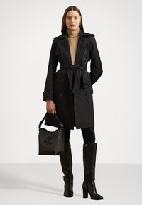 Casaco trench preto com botões duplos e cinto, combinado com botas altas pretas e uma bolsa preta e cinza com um logo.