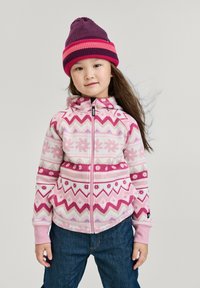 Junges Mädchen, das eine rosa gemusterte Zip-Jacke, eine lila und rosa gestreifte Mütze und eine Jeans trägt, steht vor einem schlichten Hintergrund.