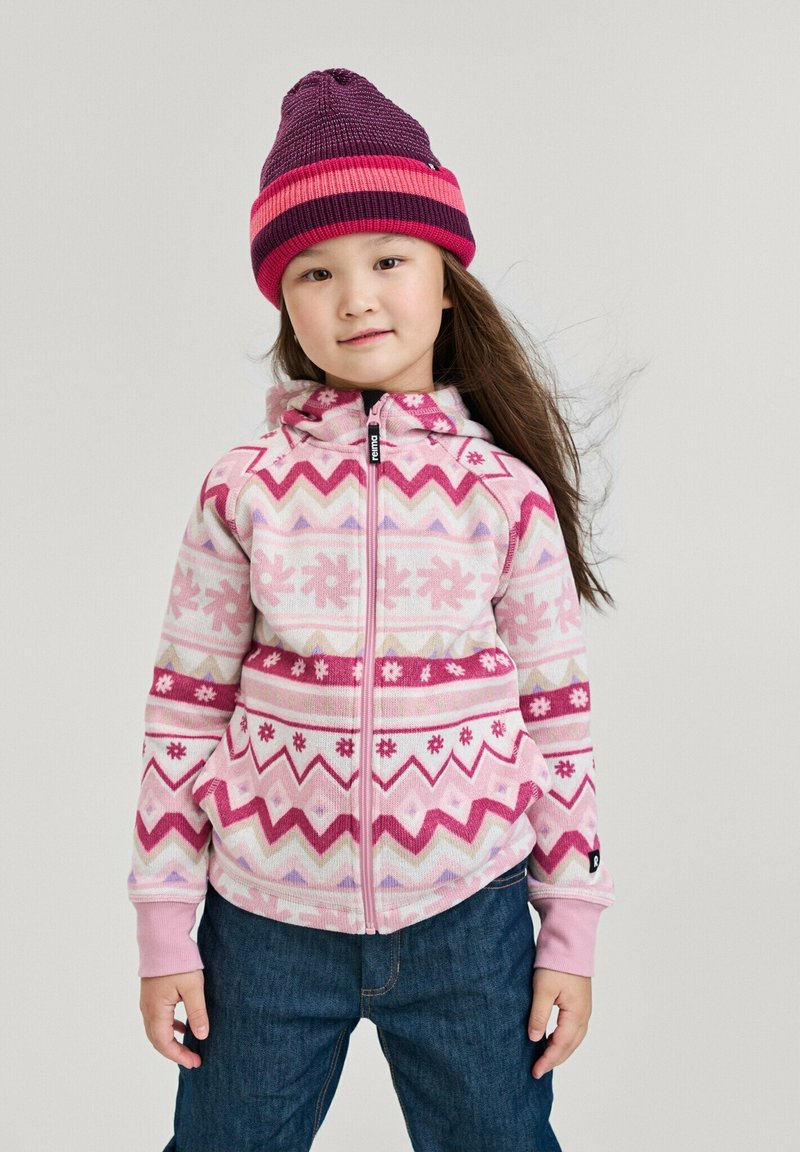 Junges Mädchen, das eine rosa gemusterte Zip-Jacke, eine lila und rosa gestreifte Mütze und eine Jeans trägt, steht vor einem schlichten Hintergrund.