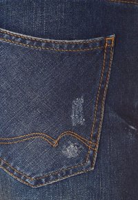 Tela de mezclilla azul oscuro con un bolsillo trasero que presenta áreas desgastadas, costuras intrincadas y un detalle de costura curvada distintivo.