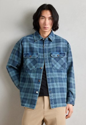 Blue plaid shirt made of denim, featuring two front pockets, brown buttons, and a classic collar. Long sleeves with cuffs.