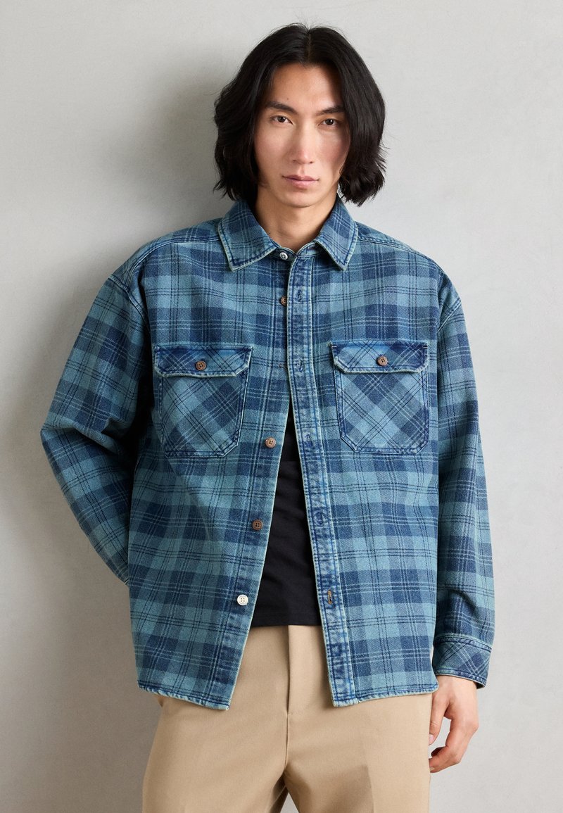 Blå rutete skjorte laget av denim, med to framlommer, brune knapper og klassisk krage. Langermet med mansjetter.