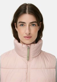 Gepolsterte Weste in sanftem Rosa, mit hohem Kragen, Reißverschluss und grünen Akzenten. Glatte Textur mit horizontalen Steppnähten.