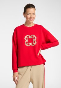 Felpa rossa dal taglio ampio, con un design di nodo in paillettes dorate sul davanti. Tessuto morbido, polsini a costine e scollo rotondo.