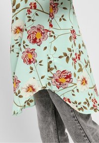 Haut à fleurs en tissu vert menthe, présentant de grandes fleurs roses et rouges avec des centres jaunes et des feuilles vertes, taillé avec un ourlet asymétrique.